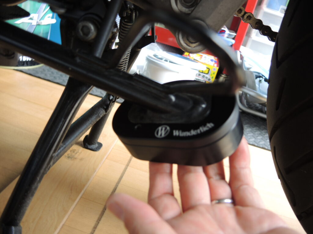 Wunderlich（ワンダーリッヒ）製のサイドスタンド拡大プレート（サイドスタンドエンラージャー）が取り付けられた状態を示す写真です。サイドスタンドの接地面にプレートを被せることで設置面積を広げ、アスファルトの熱による沈み込みや、砂利道、柔らかい地面でのバイクの転倒リスクを軽減します。このアクセサリーはBMWのG650GSなどのモデルに対応しており、バイクの駐車安定性を向上させます。