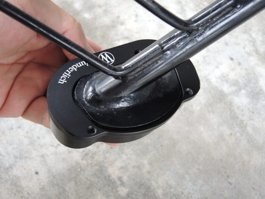 Wunderlich（ワンダーリッヒ）製のサイドスタンド拡大プレート（サイドスタンドエンラージャー）が、バイク（BMW G650GSなど）の純正サイドスタンドに装着されている近接写真です。プレートには「Wunderlich」のロゴが確認できます。このアクセサリーは、スタンドの接地面積を広げることで、柔らかい地面やアスファルトでの駐輪時の安定性を高め、バイクの転倒防止に役立ちます。