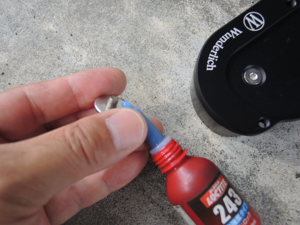 Wunderlich（ワンダーリッヒ）製のサイドスタンド拡大プレートを取り付ける際、付属のボルトにLoctite 243（ロックタイト243）中強度ネジロック剤を塗布している様子の近接写真です。指でボルトを持ち、ネジ山に青色の接着剤を少量塗布しています。この作業は、走行中の振動によるボルトの緩みを防ぎ、アクセサリーの確実な固定と安全性を高めるために行われます。