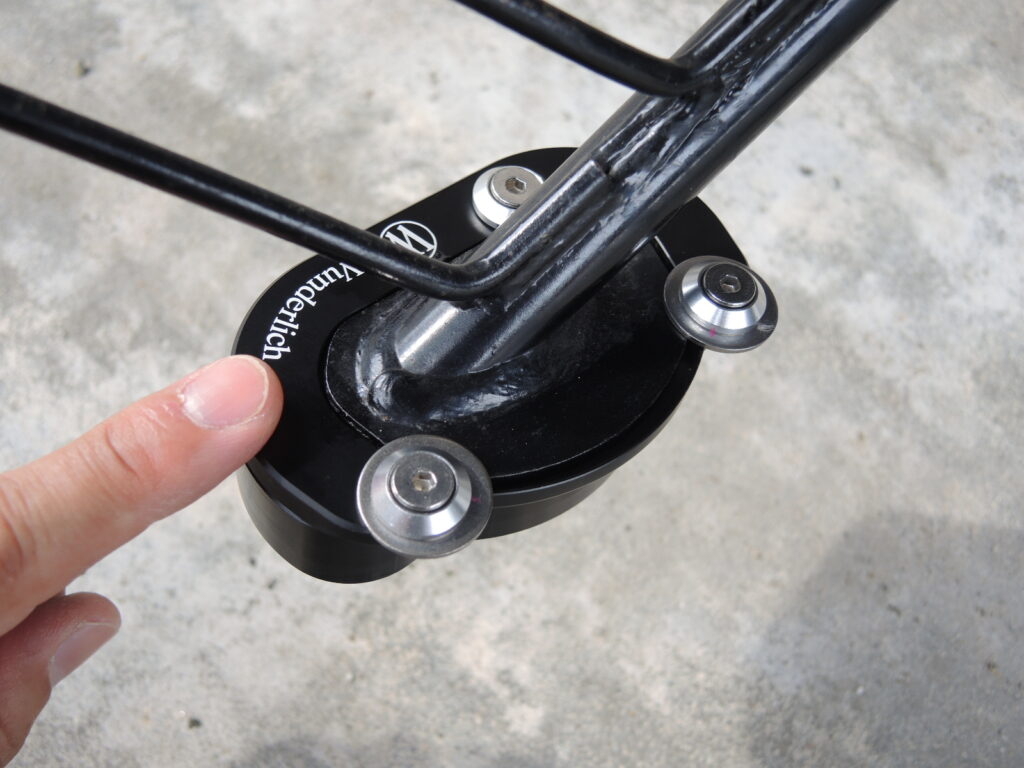 Wunderlich（ワンダーリッヒ）製のサイドスタンド拡大プレート（サイドスタンドエンラージャー）が、バイクの純正サイドスタンド底部に取り付けられた状態の近接写真です。プレートには「Wunderlich」のロゴと、それを固定する3本の六角ボルトが確認できます。手前のボルトの頭に指が添えられており、取り付け作業中であることがわかります。このアクセサリーは、BMW G650GSなどのモデルに対応し、駐輪時の安定性を高めるためのものです。