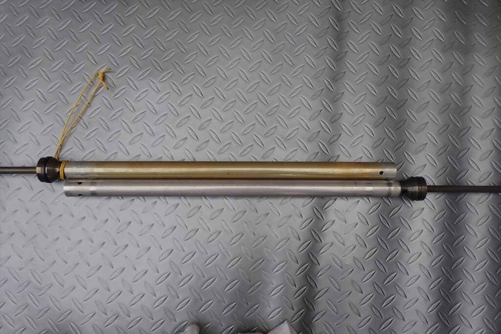 Comparing two different motorcycle suspension fluid cylinders laid out on a metal plate.