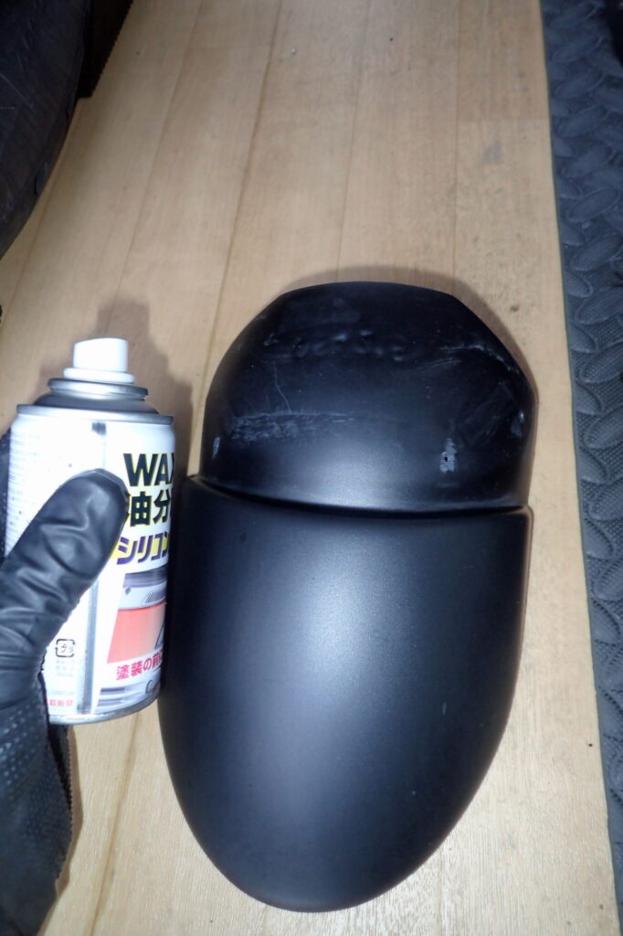 A gloved hand holding a degreaser spray can next to the bonding surface of a matte black motorcycle fender extension.
