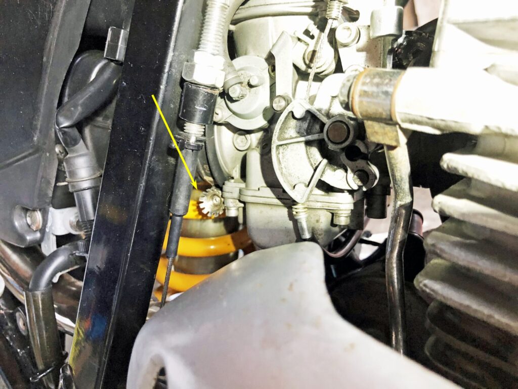 NGCスムーズドライブ(静電気除去システム)の除電ボルトが取り付けられたキャブレターとエンジン周りの詳細写真です。エンジンとフレームを繋ぐボルトや、キャブレターのエアベントホース(黄色い矢印で示されている部分)、スロットルケーブルなど、複数の箇所に施された除電対策が確認できます。