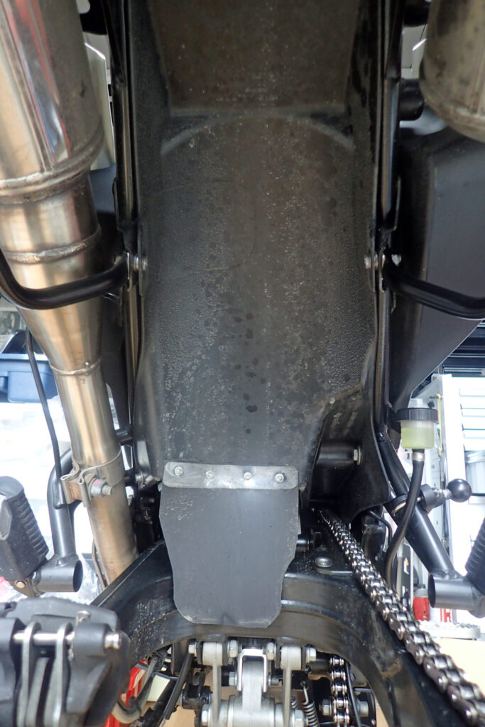 G650GSリアサスペンション下からのアングル。泥はね対策のマッドガード延長と、排気系（エキゾースト）の取り回し、スイングアームピボット周辺の整備点検ディテール。