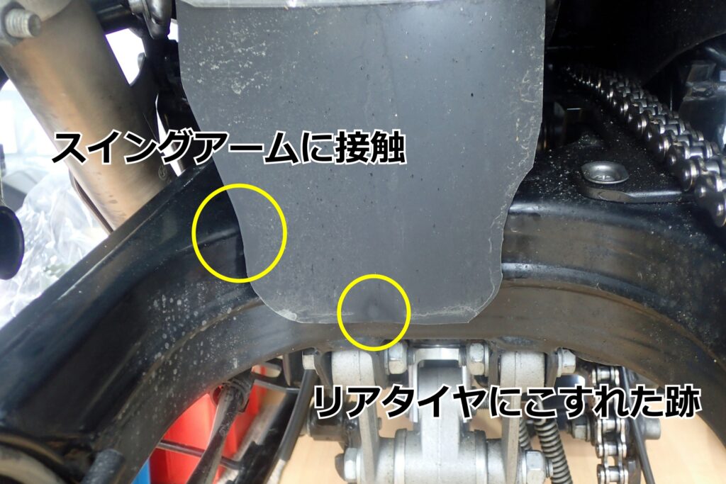 バイクのリアフェンダー延長部がスイングアームとリアタイヤに接触している箇所のクローズアップ画像。カスタムや社外品マッドガードの干渉トラブル例。