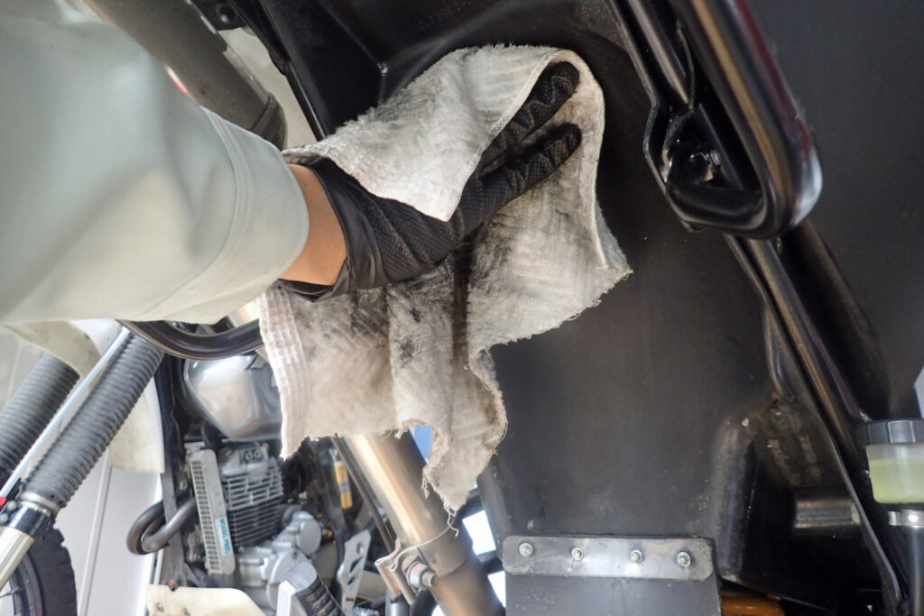 グローブを着用し、汚れたウエスでバイクの下回りを丁寧に拭き取る清掃作業。カスタムや整備前に行うパーツクリーニングの手順を示す写真。