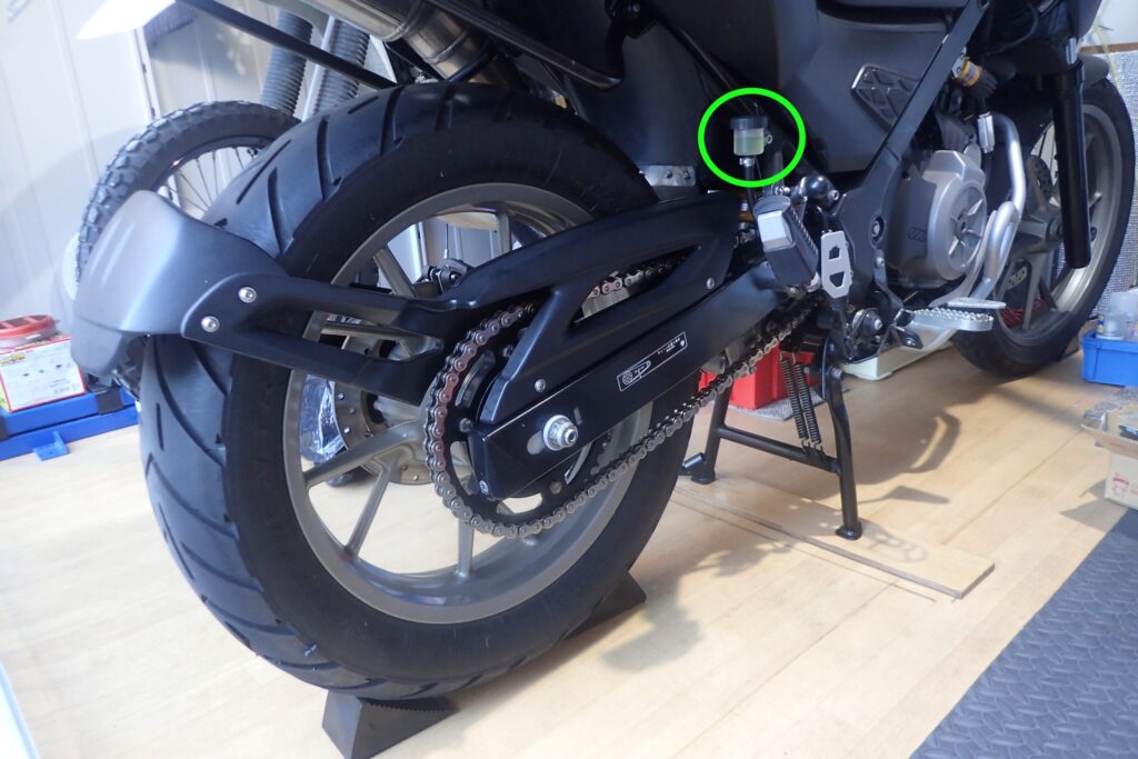 バイクのリア全体とリアブレーキリザーバータンクの位置を示す画像。後輪上部のシートレール付近に緑色の丸で囲まれたタンクが見える