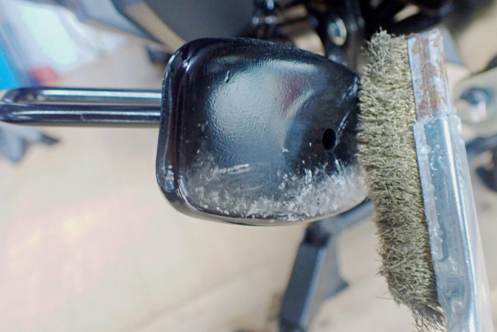Cleaning the bottom of a Honda X-ADV motorcycle side stand with a brass wire brush for rust prevention.