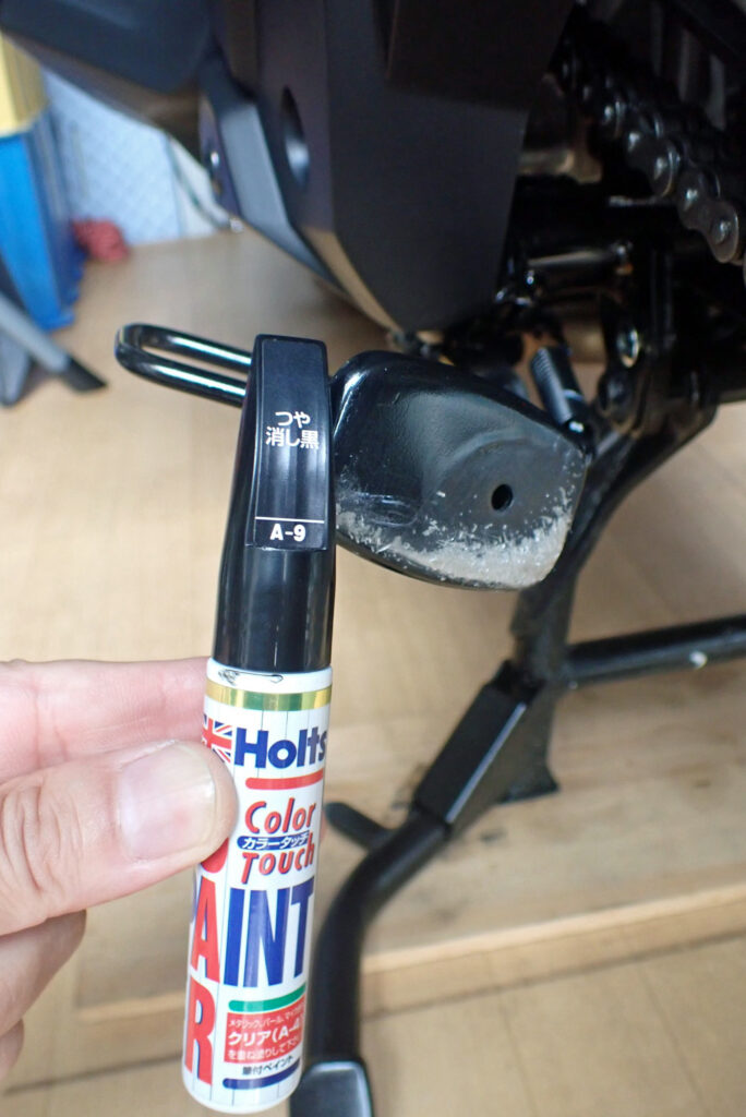 Holding a Holts matte black touch-up paint pen in front of a scratched motorcycle side stand for repair.