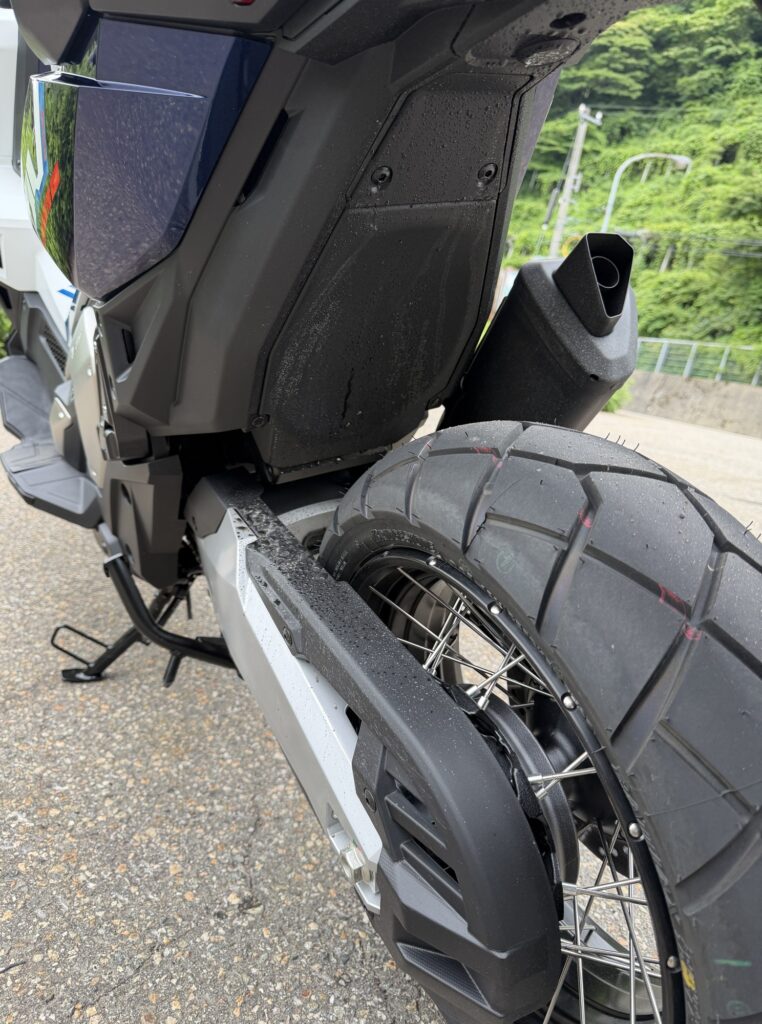 Rear left view of a Honda X-ADV 750 after riding on a wet road, showing mud splashes on the swingarm and under-tail plastic.