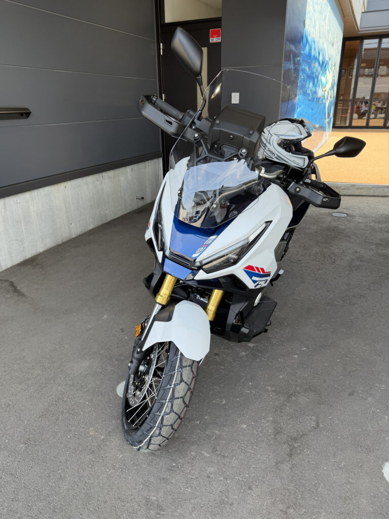 Front three-quarter view of a newly delivered Honda X-ADV 750 Special Edition motorcycle in tricolor, featuring a SHOEI full-face helmet on the seat.