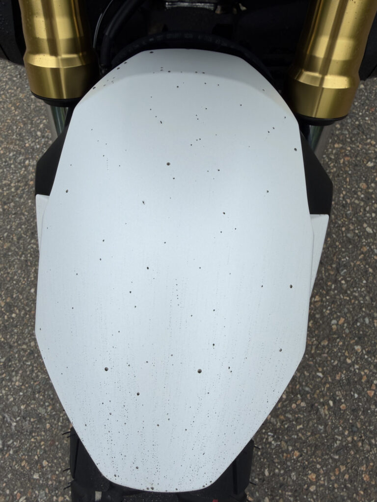 Top-down view of the white front fender on a Honda X-ADV covered with numerous black spots of mud and sand from wet road riding.