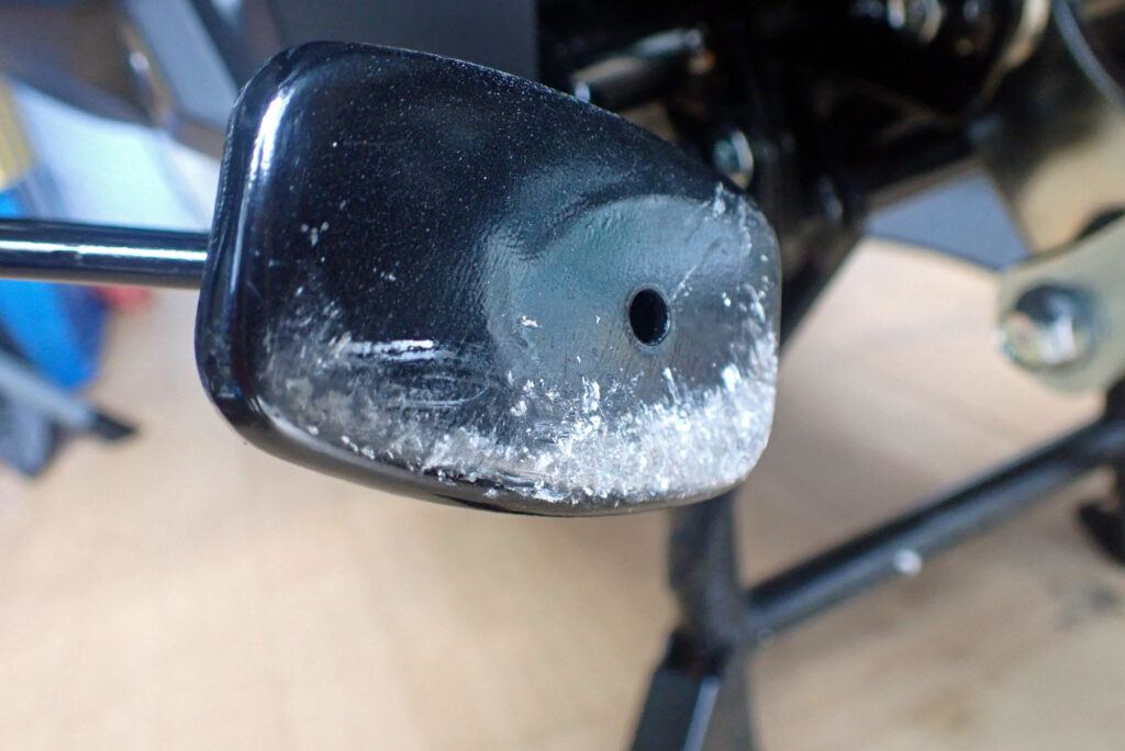 Close-up of the scratched bottom surface of a Honda X-ADV side stand, showing exposed metal with paint peeled off.