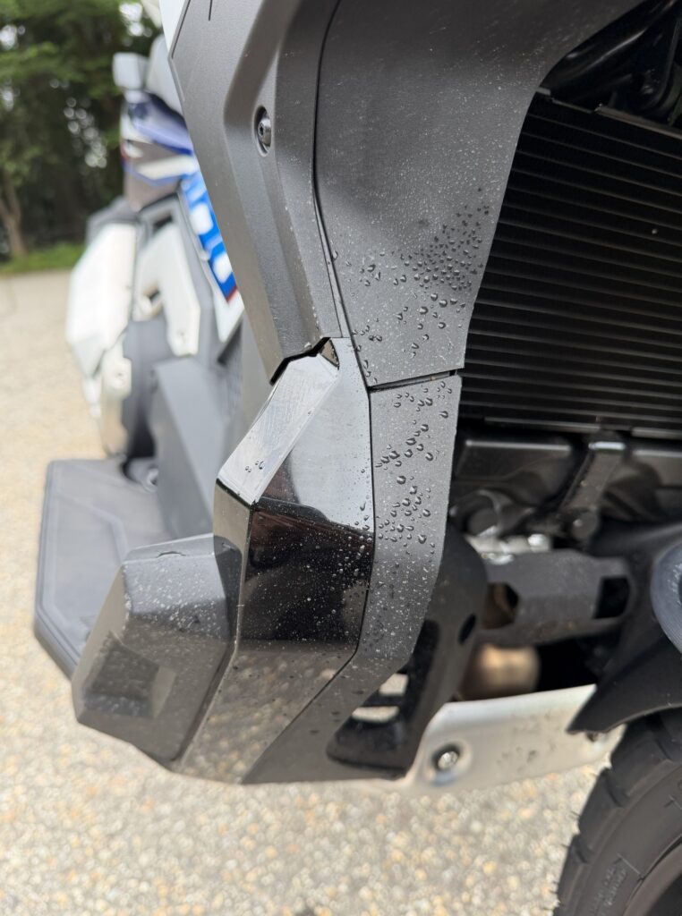 Close-up of the black side cowl and front floorboard area on a Honda X-ADV, stained with fine mud water droplets.