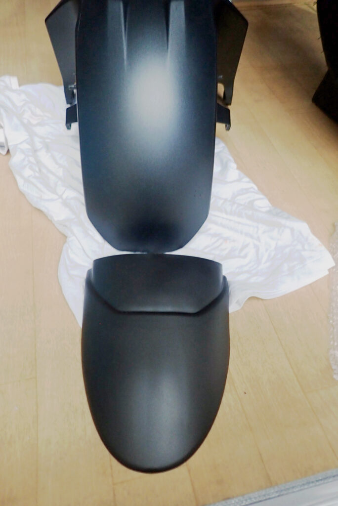 The Honda X-ADV original front fender and an unbranded matte black front fender extender extension flap laid out on the floor.