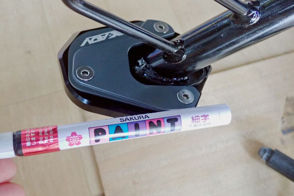 Drawing a pink alignment mark on a bolted side stand extension plate using a paint marker for loose bolt checks.
