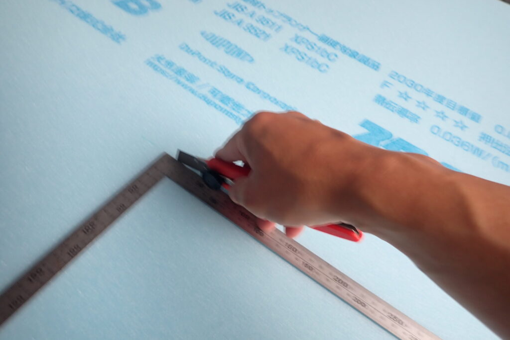 A person using a long metal ruler and a blue utility knife tool to cut a light-blue Styrofoam insulation board on the wooden floor next to a Honda XR BAJA motorcycle.