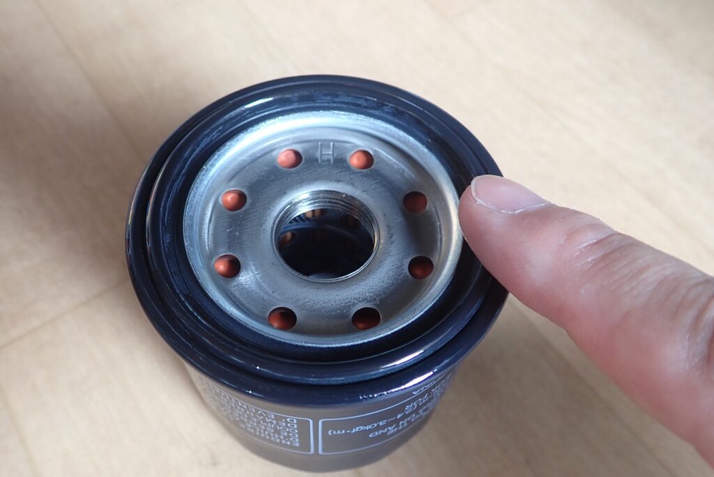 A finger applying a thin layer of fresh engine oil onto the black rubber gasket sealing ring of a new motorcycle oil filter.