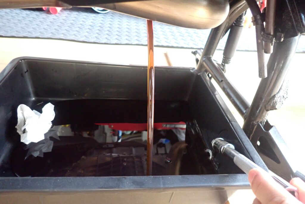 Old dark brown engine oil draining straight down into a black waste oil pan from the underdrain port of a Honda X-ADV.