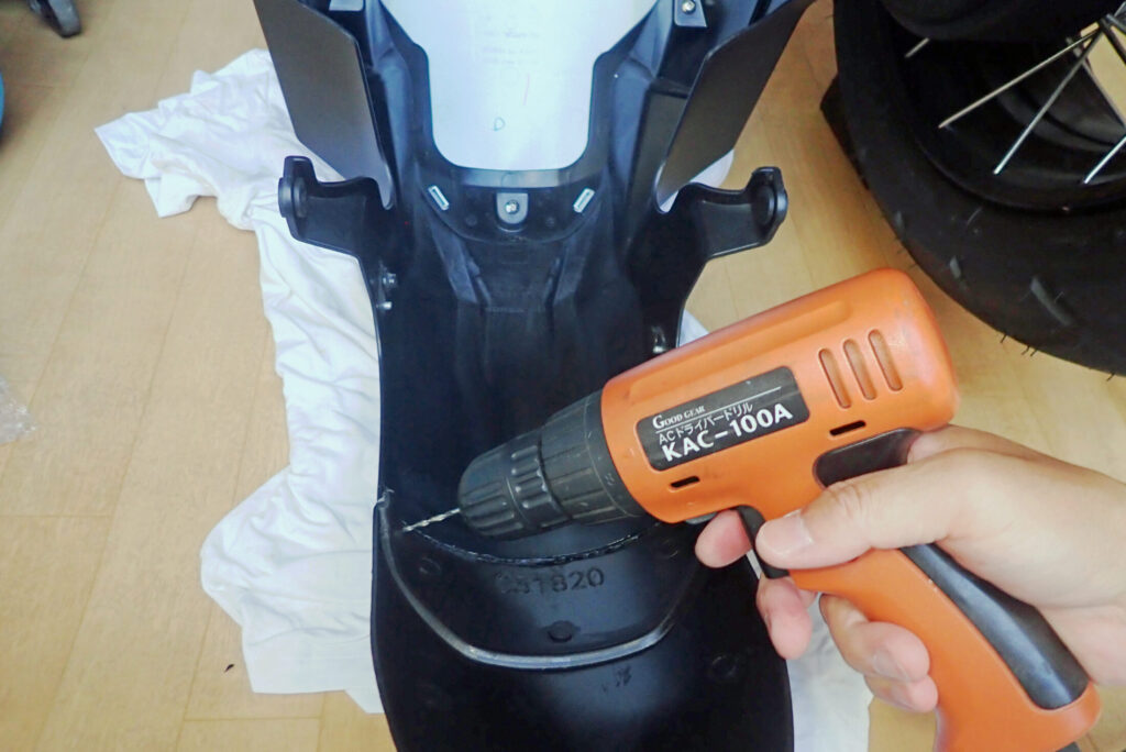 Using an orange electric drill to make guide holes on a detached motorcycle front fender for installing anti-drop safety plastic pins.
