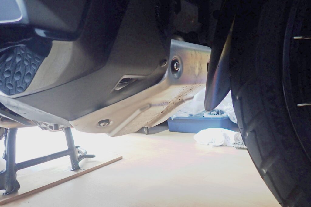 Underbody view of a Honda X-ADV 750 showing the factory aluminum skid plate underguard before removal for oil change.