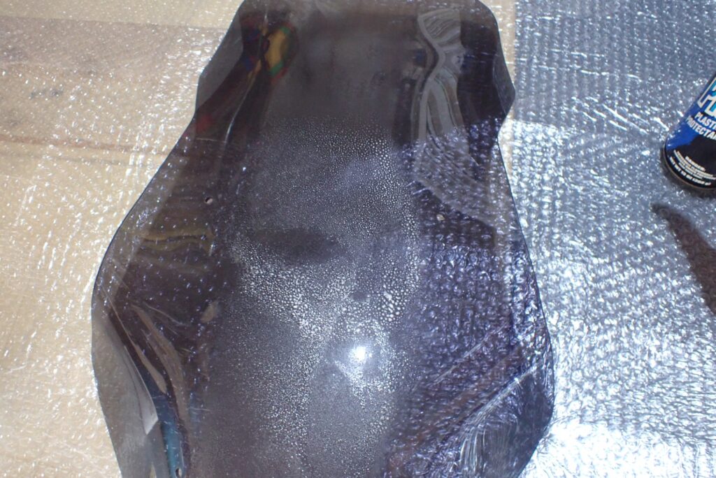 The white active foam of Plexus plastic cleaner spreading over a smoked motorcycle windscreen for high-gloss coating.