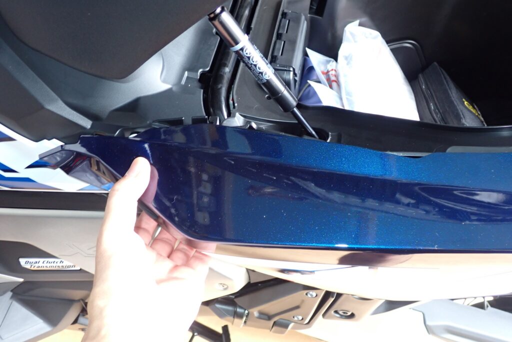 A mechanic pulling the front section of the blue Honda X-ADV rear fairing outward to release the inner mounting pins from rubber grommets.