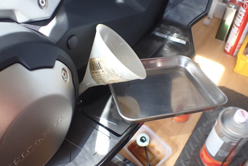 A white plastic funnel inserted into the oil filler hole on a Honda X-ADV, with a stainless steel tray placed underneath to catch any drips.