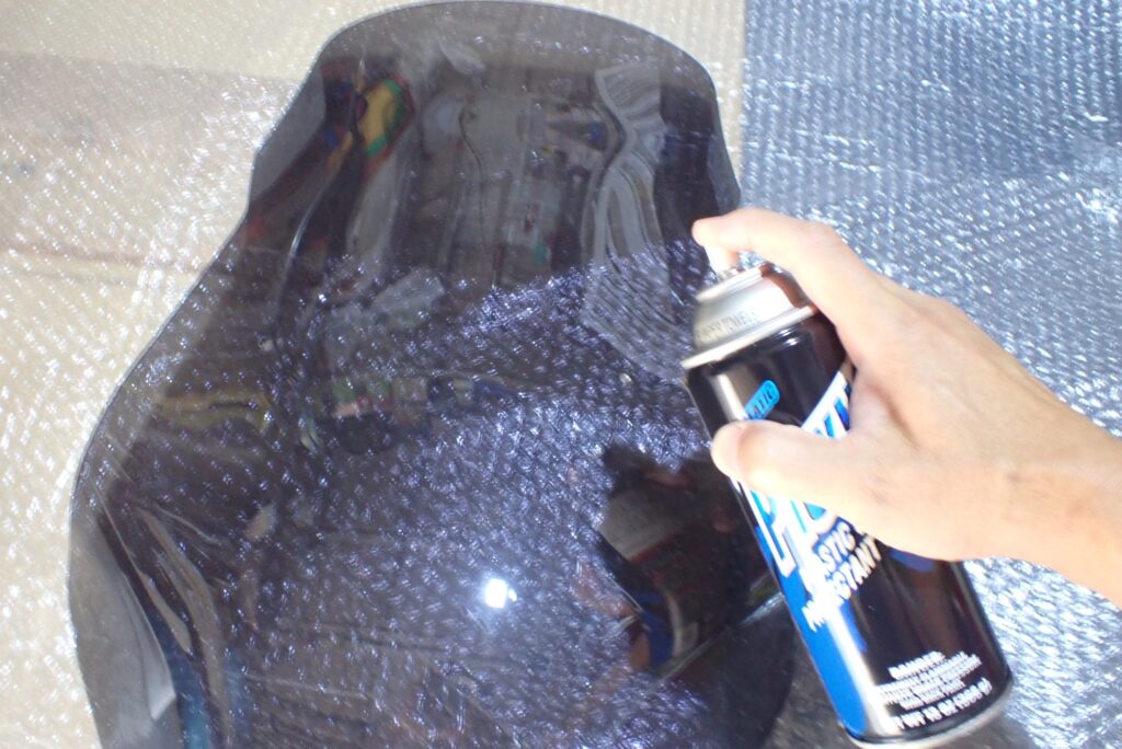 A hand spraying Plexus plastic spray directly onto the surface of a smoked motorcycle touring windscreen.