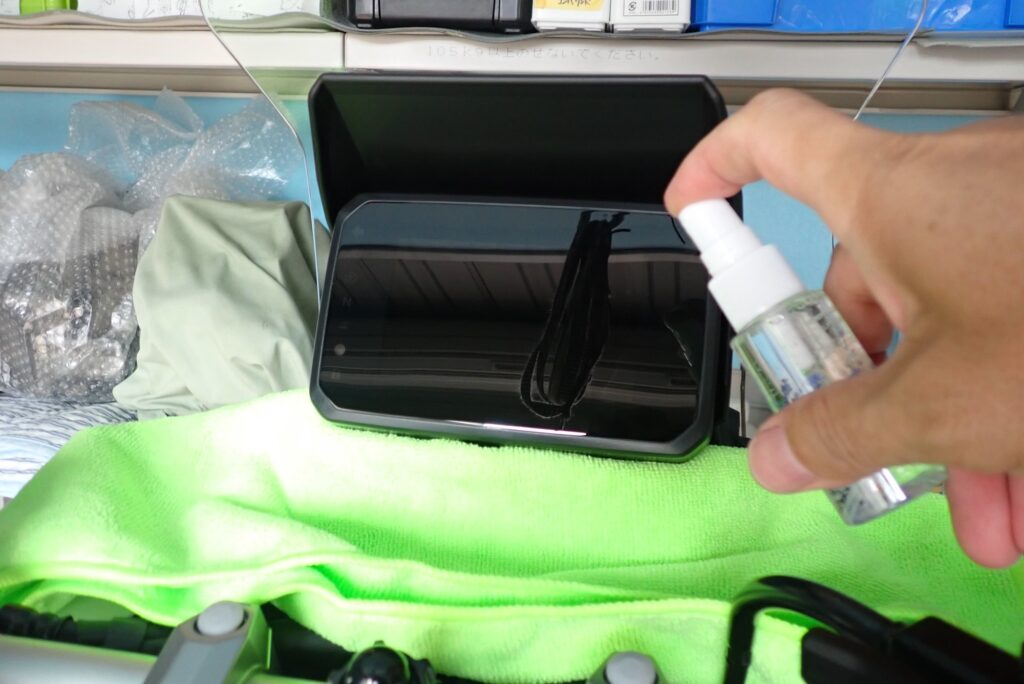 A hand spraying water mist solution onto the motorcycle TFT screen using a small bottle, with a bright green microfiber towel protecting the surrounding handle area.