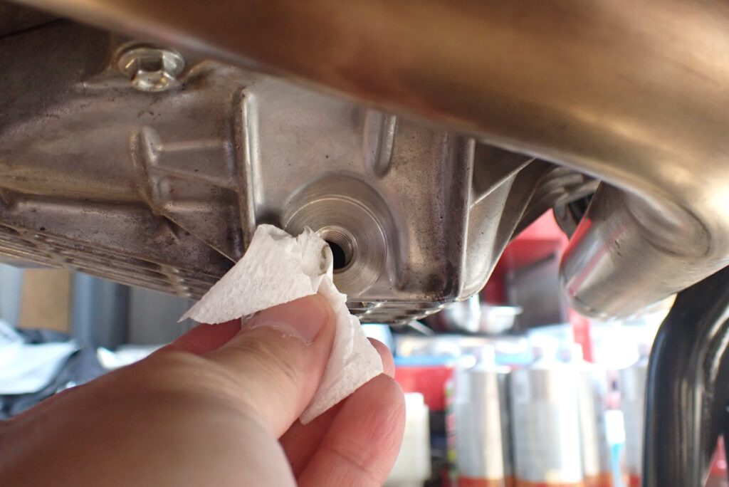 Using a clean white tissue paper wiper to clear out oil residue from the aluminum oil pan drain plug hole threads under the motorcycle.