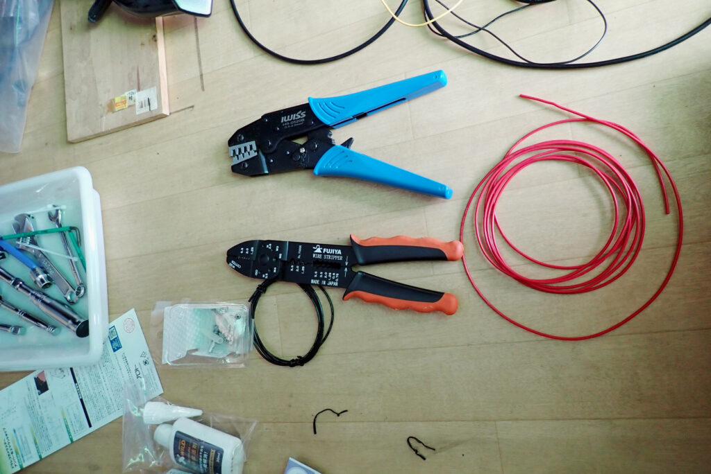 Overview of IWISS HS-0520B ratchet crimping tool, Fujiya wire stripper, and red electric wire prepared on a wooden floor.