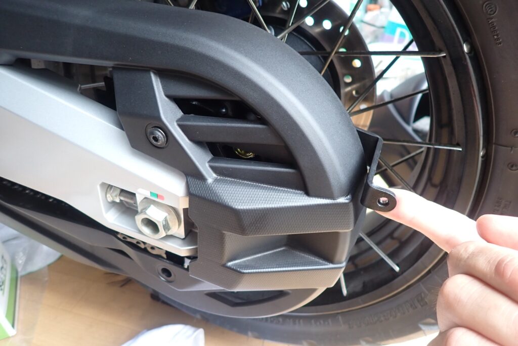 Loosening the stock chain guard to insert the Puig left-side black metal mounting bracket underneath on a Honda X-ADV.