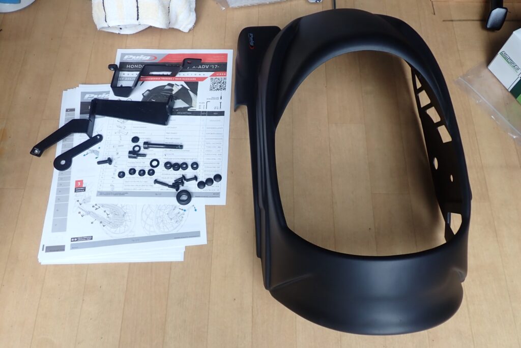 Unboxed Puig rear splash guard kit for Honda X-ADV including the black plastic hugger, metal brackets, bolts, and instruction manual.