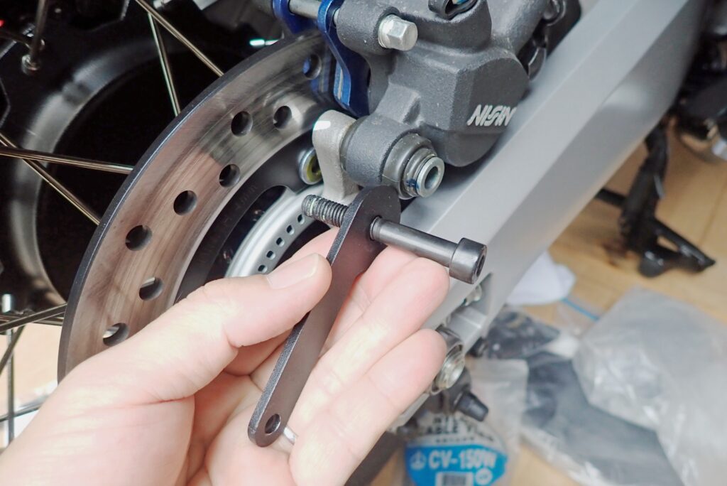 Hand holding the new Puig extended bolt and bracket next to the rear brake caliper mounting point on a Honda X-ADV.