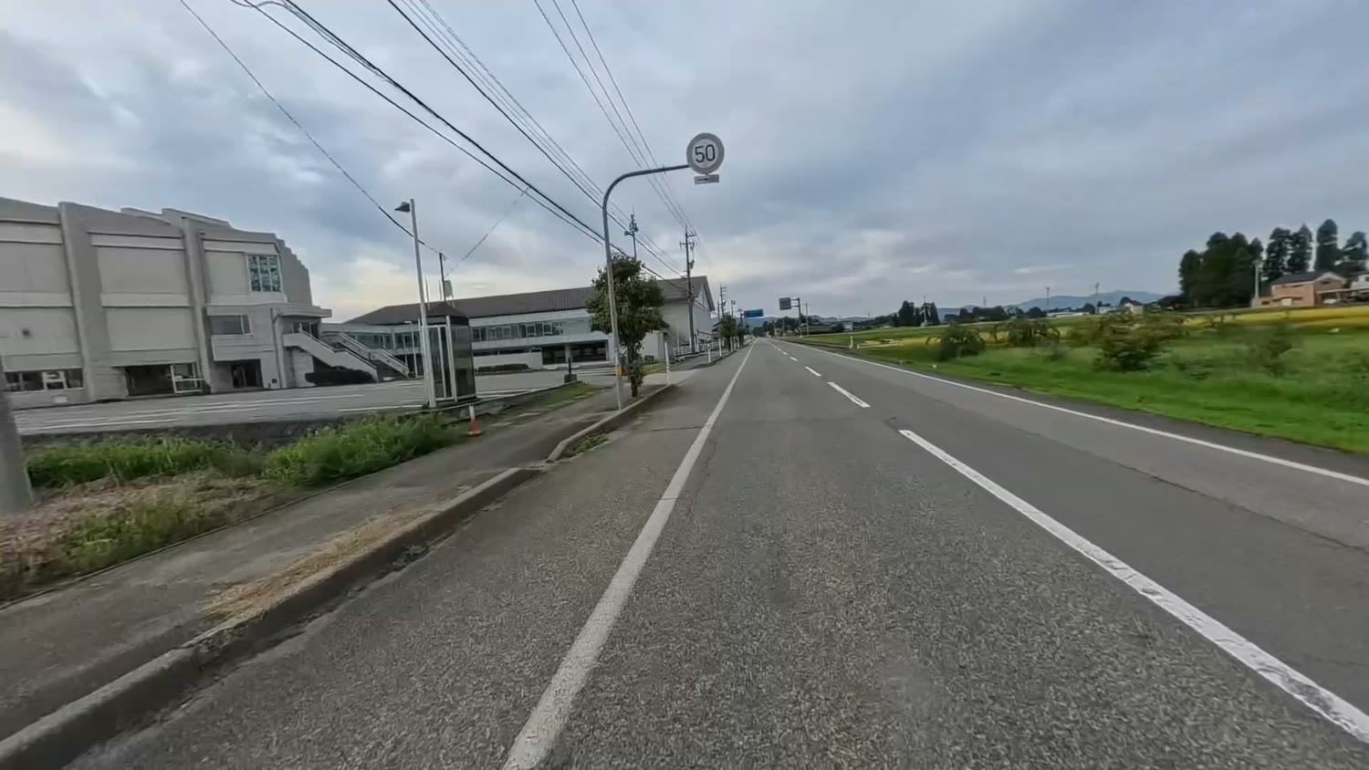 田んぼと建物のあいだをまっすぐ延びる片側1車線の道。遠くに山が見える